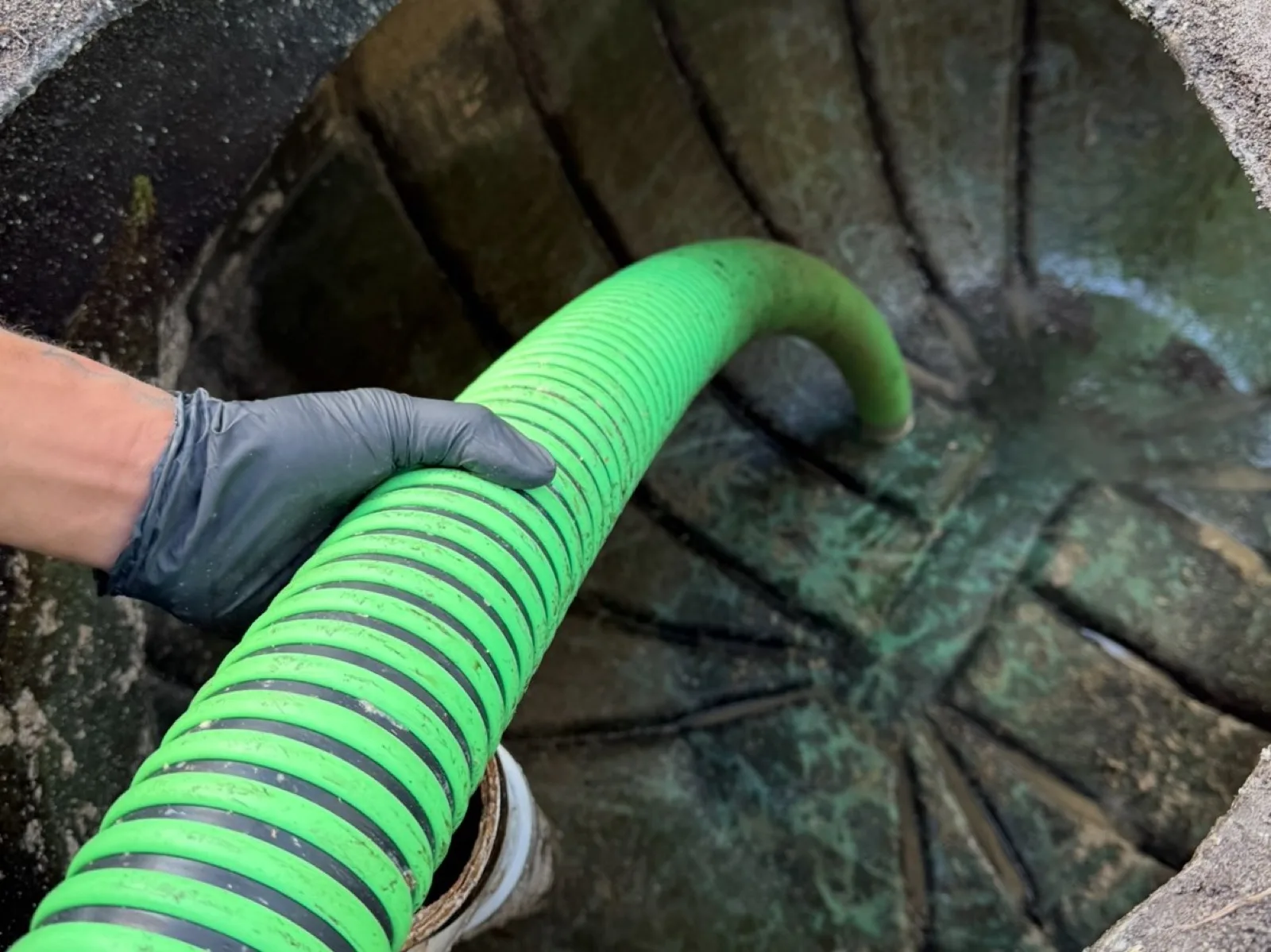 A gloved hand feeds a green vacuum hose down into an open septic tank access, with the inside of the tank visible below